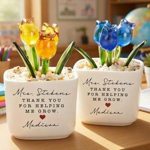 以下が含まれることがあります： ガラスのチューリップの花が入った白い正方形のプランターが2つ。1つは黄色とオレンジ色のチューリップ、もう1つは青色です。各プランターには「Mrs. Stekens, Thank you for helping me grow, Madison.」と書かれています。