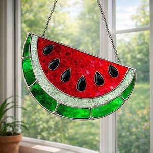 May include: A stained glass watermelon slice suncatcher. The red center has black seed details, bordered by a white, glittery rind and green outer rind. It hangs from a chain, perfect for a window decoration.