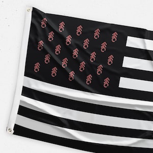 May include: Black and white flag with a design featuring horizontal stripes and a black field with a pattern of red symbols. The flag is hanging, with grommets visible on the left side.