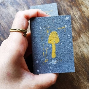 May include: A small gray card with a gold mushroom illustration. The card has a speckled texture.