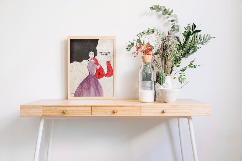 K&ouml;nnte beinhalten: Eine gerahmte Illustration einer Frau mit langen dunklen Haaren, die einen roten Fuchs h&auml;lt. Der Text "Embracing the wild soul" ist auf der Illustration geschrieben. Die gerahmte Illustration befindet sich auf einem Holztisch mit einer Blumenvase und einem Glas mit Milch.