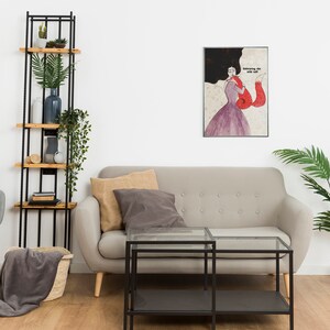 K&ouml;nnte beinhalten: Ein Wohnzimmer-Interieur mit einem grauen Sofa, zwei schwarzen Glas-Couchtischen, einem grauen Sessel, einem schwarzen Metall-Leiterregal mit Holzb&ouml;den, einer Topfpalme und einem gerahmten Druck einer Frau in einem roten Kleid mit dem Text "Knowing the wild one".