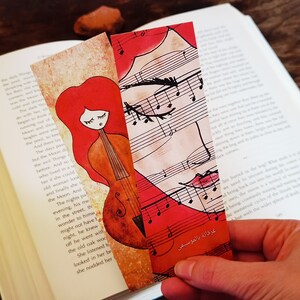 May include: Two illustrated bookmarks, one with a red-haired woman and cello, the other with a face and musical notes. The bookmarks are placed in an open book. The text "غرقانة بالموسيقى" is visible on one bookmark.