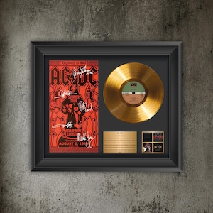 Puede incluir: Un marco que contiene un disco de vinilo dorado, un póster de concierto rojo de AC/DC del 2 de agosto de 1979 en el Riverfront Coliseum con firmas, y una placa dorada con texto sobre el álbum Back in Black y pequeñas fotos de la banda.