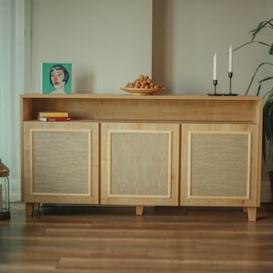 Könnte beinhalten: Ein helles Holzschrank mit drei Türen, jede mit einem gewebten Paneel. Der Schrank hat ein Regal darüber mit einem Gemälde, Büchern und einer Schale mit Obst. Zwei Kerzenständer und eine Topfpflanze sind ebenfalls sichtbar.