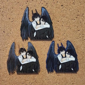 Pode incluir: Três pinos de esmalte com um casal abraçado, cada um com grandes asas pretas. As figuras usam ternos pretos e camisas brancas. Uma figura tem chifres. Os pinos estão sobre um fundo castanho texturizado.