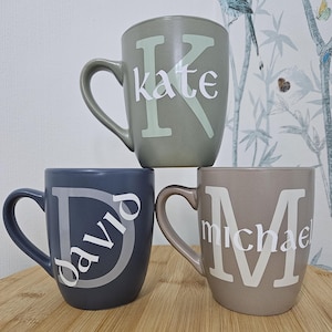Puede incluir: Tres tazas de cerámica con nombres personalizados. Las tazas están apiladas una encima de la otra. La taza de arriba es verde con el nombre "kate" en letras blancas. La taza del medio es azul con el nombre "david" en letras blancas. La taza de abajo es marrón con el nombre "michael" en letras blancas.