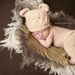 Puede incluir: Un bebé recién nacido con un gorro de oso de crochet y pantalones a juego, durmiendo en una manta de textura marrón y blanca.