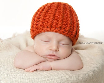 May include: A sleeping baby wearing an orange crocheted hat, lying on a white blanket.