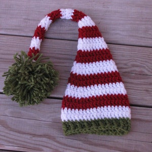 Puede incluir: Un gorro de elfo de rayas rojas y blancas con un pompón verde en la parte superior. El gorro está tejido a crochet y tiene un ribete verde.