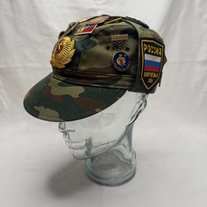 May include: Camouflage baseball cap with multiple pins and patches. The cap features a Russian flag patch and a gold emblem. The cap is displayed on a clear glass head form.