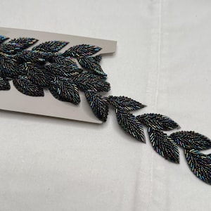 May include: A beaded leaf trim in shades of black and iridescent colors. The leaves are detailed with small beads, creating a textured surface. The trim is attached to a piece of gray paper.