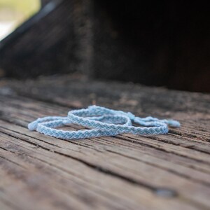 May include: Two light blue and white woven bracelets are displayed on a weathered wooden surface. The bracelets have a chevron pattern and are neatly arranged. The background is blurred, suggesting an outdoor setting. The bracelets are a simple, yet stylish accessory.