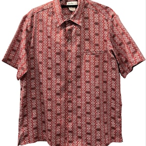 May include: A short-sleeve button-down shirt in a deep red hue featuring a repeating geometric pattern in white, black, and dark gray. The shirt includes a standard collar, a single chest pocket on the left side, and a row of matching buttons.