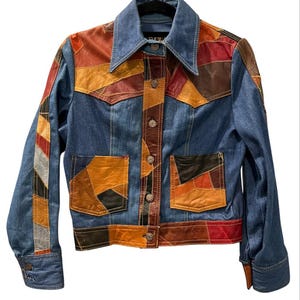 May include: A blue denim jacket featuring a patchwork design of multi-colored leather panels in shades of brown, red, and black. The jacket includes a pointed collar, a front button closure with metallic buttons, and two front patch pockets.