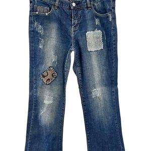 以下が含まれることがあります： ダメージ加工と装飾パッチが施されたブルーデニムジーンズ。ジーンズは、クラシックな5ポケットデザイン、ボタンとジッパーの開閉、わずかにブーツカットのレッグオープニングが特徴です。装飾には、フロントポケットとパッチのラインストーンアクセントが含まれています。