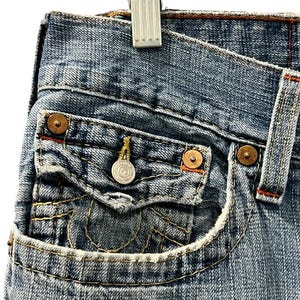 May include: Blue denim jeans featuring a small coin pocket with a flap closure, secured by a silver-toned metal button. The pocket and seams are accented with thick, contrasting yellow stitching and copper-colored metal rivets at the corners.