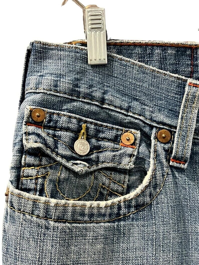 May include: Blue denim jeans featuring a small coin pocket with a flap closure, secured by a silver-toned metal button. The pocket and seams are accented with thick, contrasting yellow stitching and copper-colored metal rivets at the corners.