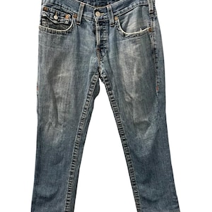 May include: Medium-wash blue denim jeans featuring a classic five-pocket design, a button fly closure, and prominent thick contrast stitching along the seams. The fabric displays a faded, worn-in texture with whiskering details at the hips.