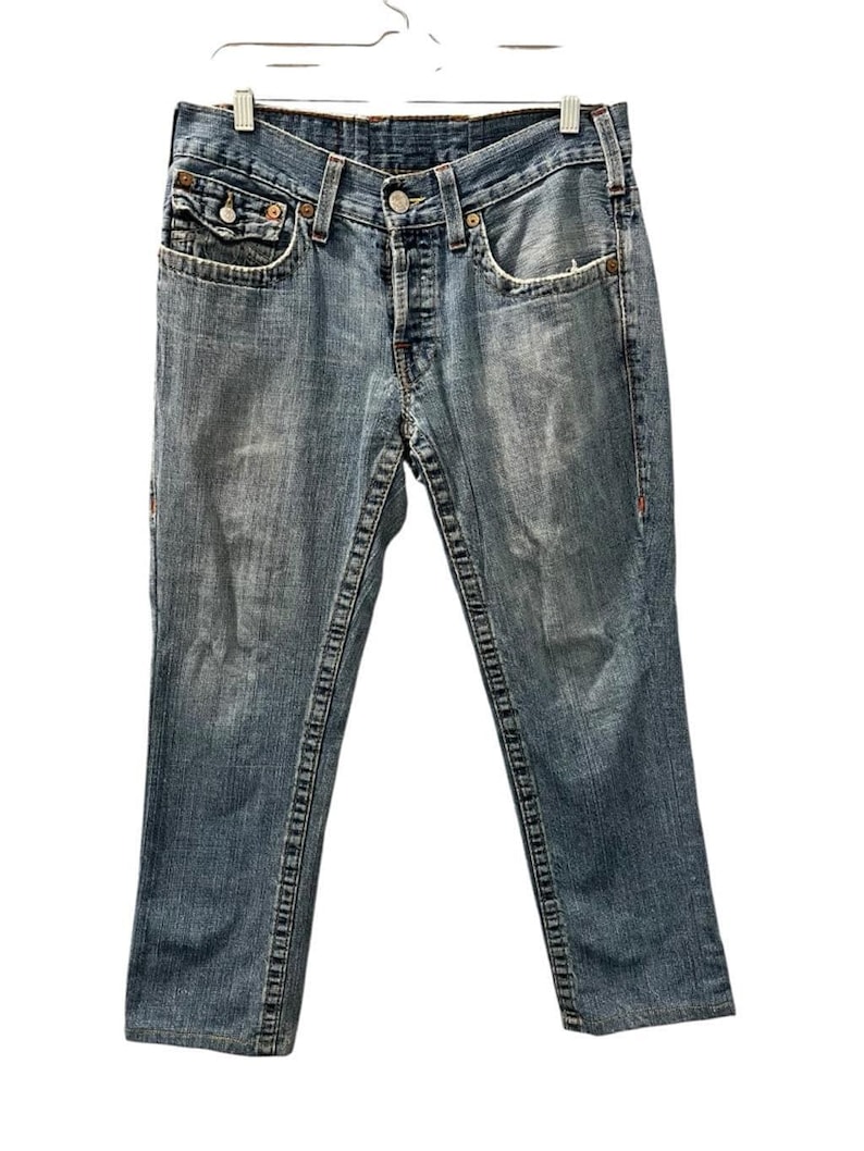 May include: Medium-wash blue denim jeans featuring a classic five-pocket design, a button fly closure, and prominent thick contrast stitching along the seams. The fabric displays a faded, worn-in texture with whiskering details at the hips.