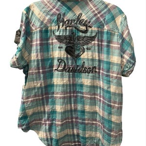 Może przedstawiać: Koszula w kratę z krótkim rękawem w odcieniach turkusu, fioletu i kremu. Z tyłu haftowany napis "Harley Davidson" z emblematem skrzydlatego serca. Koszula ma luźny krój.