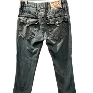May include: A pair of grey denim jeans featuring a faded wash, two back pockets with button-down flaps, thick contrast stitching, belt loops, and a tan leather brand patch on the waistband that reads True Religion with a Buddha logo graphic.