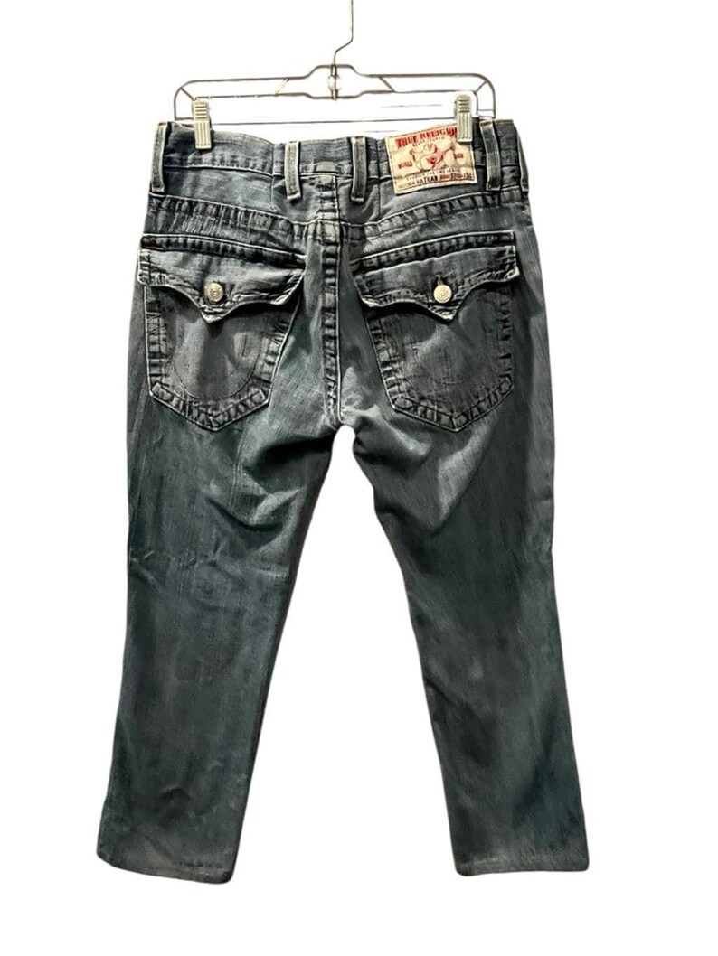 May include: A pair of grey denim jeans featuring a faded wash, two back pockets with button-down flaps, thick contrast stitching, belt loops, and a tan leather brand patch on the waistband that reads True Religion with a Buddha logo graphic.