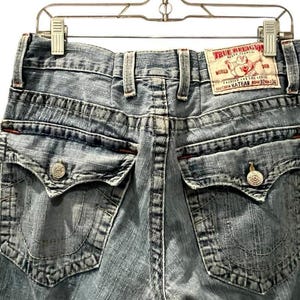 May include: Light blue denim jeans featuring a back view with two flap pockets, each secured with a silver button. A white leather patch on the waistband displays the red text True Religion Brand Jeans, a Buddha logo, and the size 32x34.