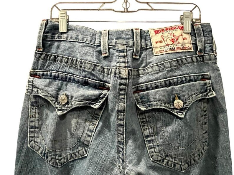 May include: Light blue denim jeans featuring a back view with two flap pockets, each secured with a silver button. A white leather patch on the waistband displays the red text True Religion Brand Jeans, a Buddha logo, and the size 32x34.