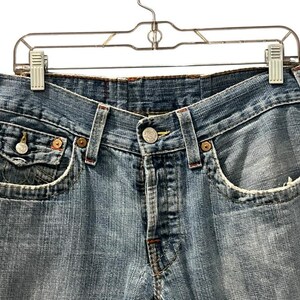 May include: Light blue denim jeans featuring a classic five-pocket design with copper-toned rivets, a button fly closure, and a small coin pocket with a buttoned flap. The fabric displays a faded wash with frayed edges along the pocket openings.