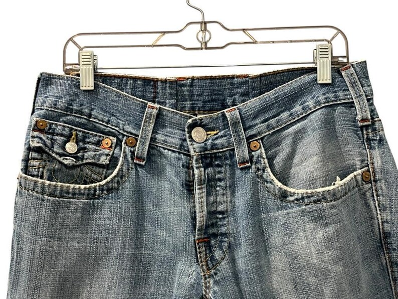 May include: Light blue denim jeans featuring a classic five-pocket design with copper-toned rivets, a button fly closure, and a small coin pocket with a buttoned flap. The fabric displays a faded wash with frayed edges along the pocket openings.
