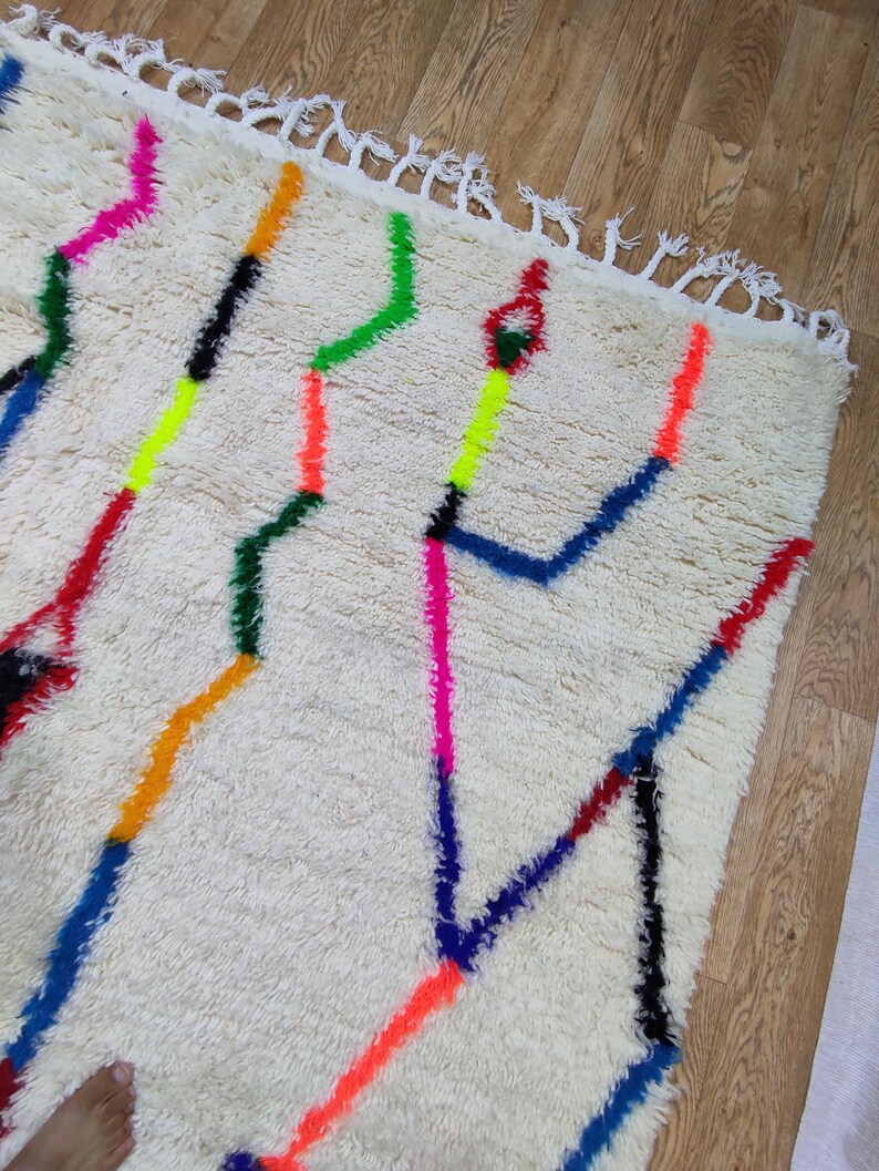 K&ouml;nnte beinhalten: Cremefarbener, strukturierter Teppich mit geometrischem Design. Der Teppich hat farbenfrohe Zickzacklinien in Neonrosa, Orange, Gr&uuml;n, Gelb, Blau und Rot. Die Kanten sind mit wei&szlig;en Fransen versehen.