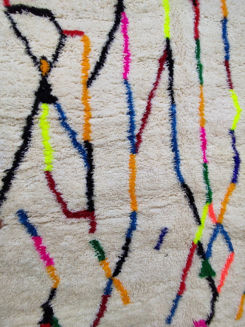 K&ouml;nnte beinhalten: Cremefarbener Teppich mit strukturierter Oberfl&auml;che und einem lebendigen geometrischen Muster. Das Design zeigt schwarze, rote, gelbe, blaue, orange, gr&uuml;ne und rosa Linien und Formen, die eine farbenfrohe und moderne &Auml;sthetik schaffen.