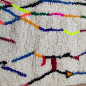 K&ouml;nnte beinhalten: Cremefarbener, strukturierter Teppich mit einem farbenfrohen, abstrakten Design. Der Teppich hat Linien in Schwarz, Blau, Rot, Orange, Gelb, Gr&uuml;n und Pink. Der Teppich hat ein weiches, pl&uuml;schiges Aussehen.