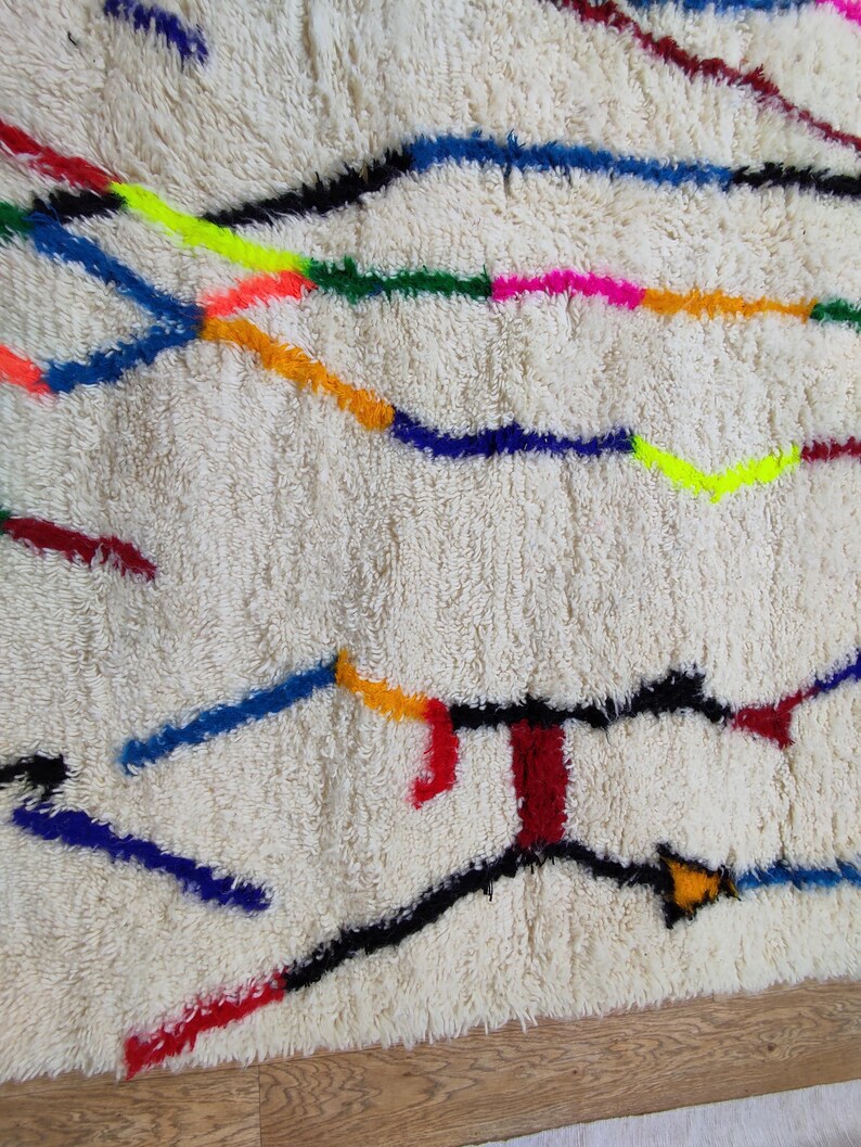 K&ouml;nnte beinhalten: Cremefarbener, strukturierter Teppich mit einem farbenfrohen, abstrakten Design. Der Teppich hat Linien in Schwarz, Blau, Rot, Orange, Gelb, Gr&uuml;n und Pink. Der Teppich hat ein weiches, pl&uuml;schiges Aussehen.