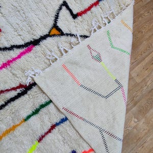 K&ouml;nnte beinhalten: Ein cremefarbener, strukturierter Teppich mit geometrischem Design. Der Teppich hat schwarze, rote, gelbe, rosa, blaue und gr&uuml;ne Linien und Formen. Der Rand des Teppichs ist sichtbar und zeigt eine gewebte R&uuml;ckseite mit farbigen Linien.