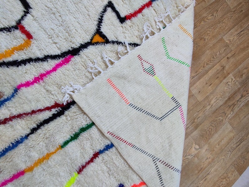 K&ouml;nnte beinhalten: Ein cremefarbener, strukturierter Teppich mit geometrischem Design. Der Teppich hat schwarze, rote, gelbe, rosa, blaue und gr&uuml;ne Linien und Formen. Der Rand des Teppichs ist sichtbar und zeigt eine gewebte R&uuml;ckseite mit farbigen Linien.