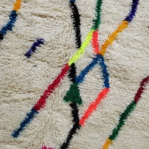 K&ouml;nnte beinhalten: Nahaufnahme eines cremefarbenen Teppichs mit zotteliger Textur. Der Teppich hat ein Design aus bunten, unregelm&auml;&szlig;igen Linien in Blau-, Rot-, Gr&uuml;n-, Gelb-, Orange- und Schwarzt&ouml;nen. Die Linien schneiden sich und &uuml;berlappen sich und erzeugen ein lebendiges Muster.