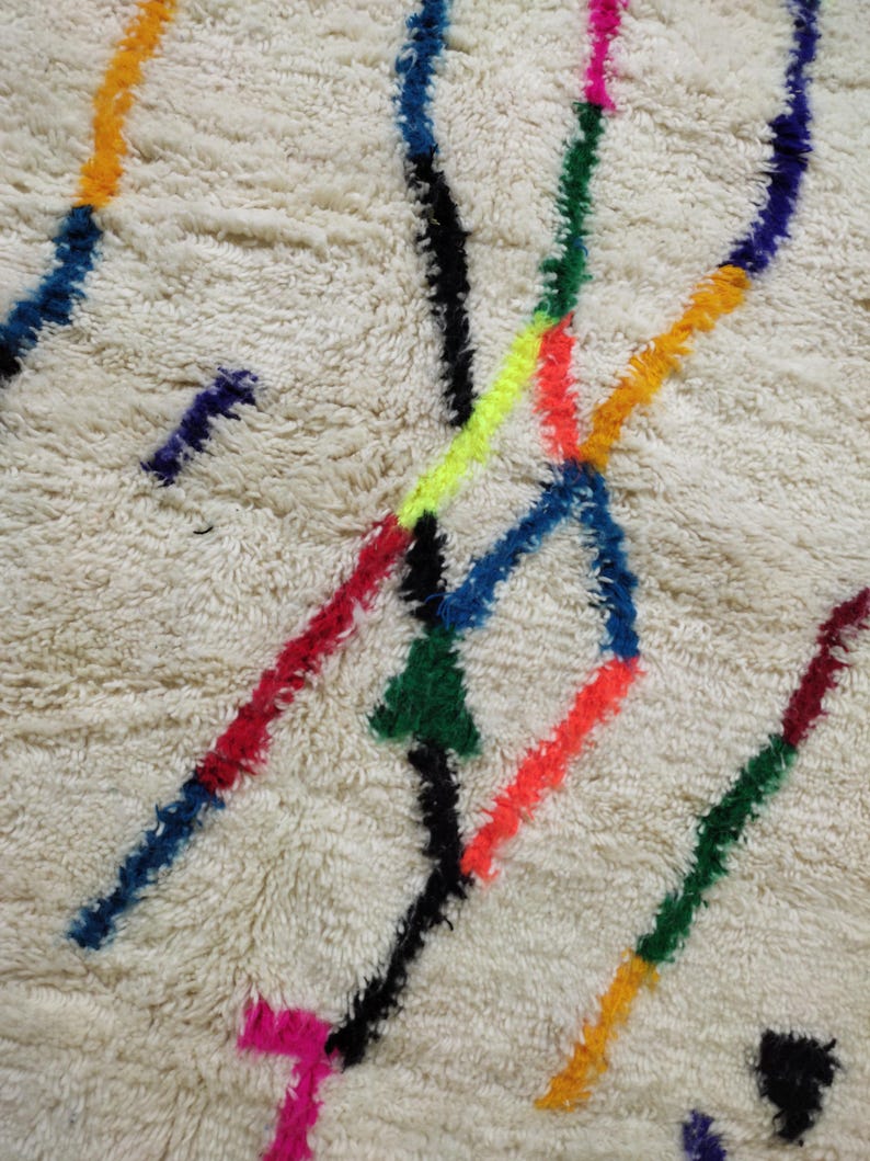 K&ouml;nnte beinhalten: Nahaufnahme eines cremefarbenen Teppichs mit zotteliger Textur. Der Teppich hat ein Design aus bunten, unregelm&auml;&szlig;igen Linien in Blau-, Rot-, Gr&uuml;n-, Gelb-, Orange- und Schwarzt&ouml;nen. Die Linien schneiden sich und &uuml;berlappen sich und erzeugen ein lebendiges Muster.