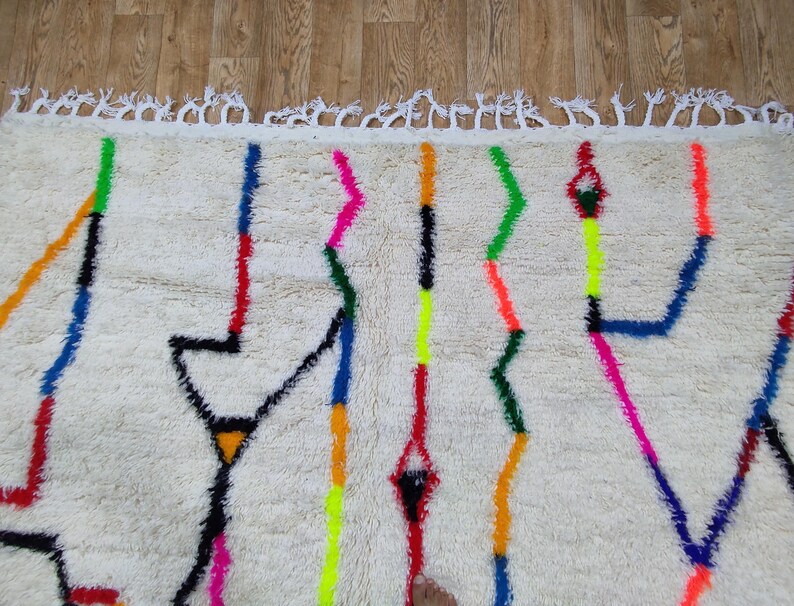 K&ouml;nnte beinhalten: Cremefarbener Teppich mit einem geometrischen Design mit leuchtenden Linien in Blau, Rot, Gelb, Gr&uuml;n und Pink. Der Teppich hat eine strukturierte Oberfl&auml;che und einen Fransenrand.