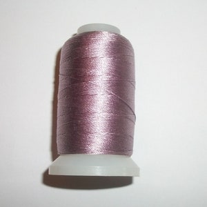 May include: A spool of mauve-colored thread. The thread is tightly wound around a cylindrical core with a white plastic base. The thread has a slight sheen, suggesting a silk or satin material. Ideal for sewing, embroidery, or crafting projects.