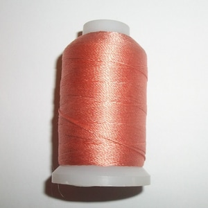 May include: A spool of coral-colored thread. The thread is tightly wound around a white plastic spool. The thread has a slight sheen, suggesting a silk or satin material. Ideal for sewing, embroidery, or other craft projects.