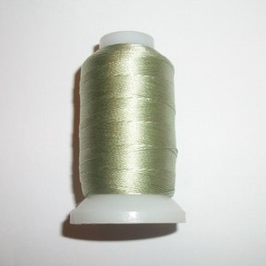 May include: A spool of olive green thread. The thread is tightly wound around a white plastic spool. The thread has a slight sheen, suggesting a silk or satin material. The spool is set against a plain white background.
