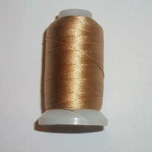 May include: A spool of metallic gold thread, tightly wound around a white plastic base. The thread has a subtle sheen, reflecting light. Ideal for sewing, embroidery, or crafting projects.