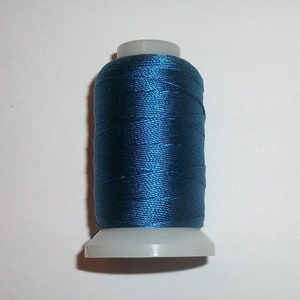 May include: A spool of dark teal thread. The thread is tightly wound around a gray plastic spool. The thread has a slight sheen, suggesting a silk or satin material. The spool is set against a plain white background.