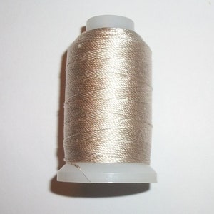 Op de afbeelding: Een spoel lichtbruin garen. De draad is strak gewikkeld rond een witte plastic spoel. De draad heeft een lichte glans, wat wijst op een zijde- of satijnachtig materiaal. De spoel staat op een wit oppervlak.