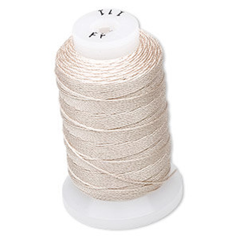 Op de afbeelding: Een spoel lichtbeige garen. De draad is strak gewikkeld rond een witte plastic spoel. De bovenkant van de spoel heeft kleine markeringen. De draad lijkt een fijn, glad materiaal te zijn, geschikt voor naai- of borduurprojecten.