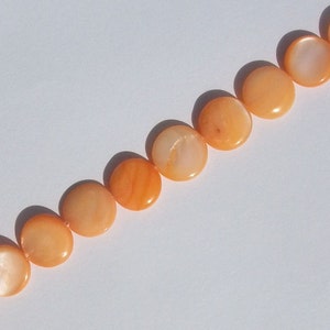 May include: A strand of orange-colored, flat, round beads. The beads have a glossy finish and are arranged diagonally across a white background. The beads are uniform in size and shape, suggesting they are for jewelry making.