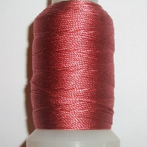 May include: A spool of red-pink thread. The thread is tightly wound around a white plastic spool. The thread has a slight sheen, suggesting a smooth texture. The spool is upright against a white background. This thread is likely for sewing or embroidery projects.