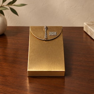 May include: A rectangular gold-toned metal case with a semi-circular flap closure. The flap features a silver-colored Space Needle emblem and a small square plaque with the text SEATTLE WASHINGTON 1962.